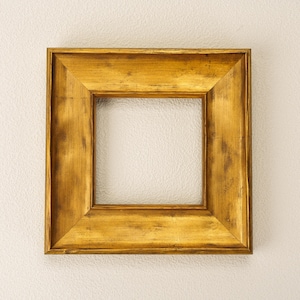 May include: Square, gold-colored picture frame with a distressed finish. The frame has a thick, beveled edge and a warm, antique appearance. The frame is empty, ready to hold a photograph or artwork.