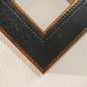 Puede incluir: Primer plano de una esquina de un marco de fotos negro con borde dorado. El marco tiene una apariencia texturizada, similar a la madera. El borde dorado presenta un patrón triangular repetido, agregando un elemento decorativo al diseño del marco.