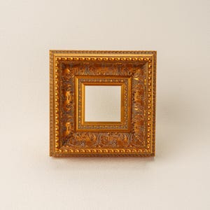 Puede incluir: Un marco de fotos cuadrado y dorado con un diseño detallado y texturizado. El marco tiene una abertura cuadrada más pequeña en el centro. El marco es probablemente para exhibir una fotografía o una obra de arte.