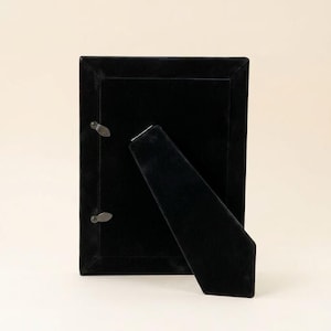 May include: A rectangular black velvet picture frame shown from the back. The frame features a matching black velvet easel stand attached to the centre and two small metal swivel clips on the left side to secure the backing in place.
