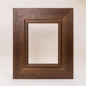 Puede incluir: Un marco de fotos cuadrado de madera con acabado marrón oscuro. El marco tiene un diseño en capas con un borde interior empotrado. La veta de la madera es visible, agregando textura al marco.