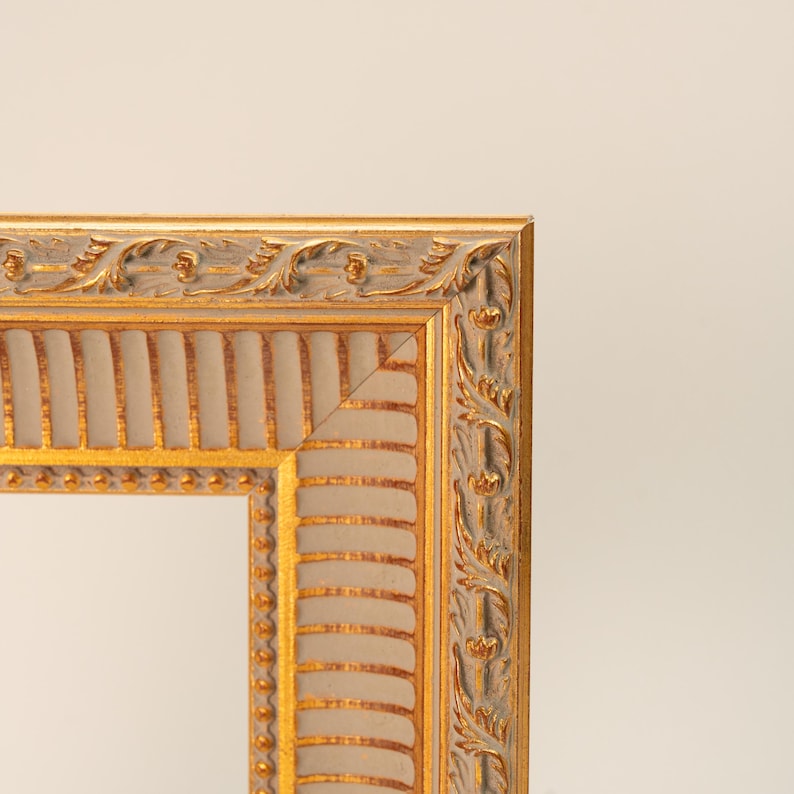 May include: Close-up of a decorative picture frame with a gold and silver finish. The frame has ornate detailing along the edges, with a repeating pattern of leaves and floral elements. The inner portion of the frame is a light beige color.