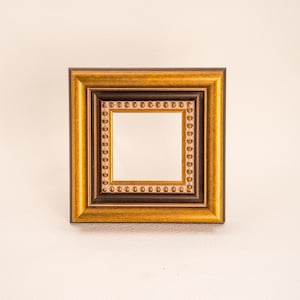 May include: A square picture frame featuring a wide, textured gold-toned outer border, a dark brown inner trim, and a central square section decorated with a row of small, raised gold-colored studs surrounding a plain, light-colored center.