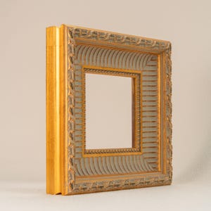 May include: Ornate gold picture frame with a light gray inner border. The frame has detailed carvings and a square opening for a photo or artwork. The frame is designed to be displayed on a wall or tabletop.