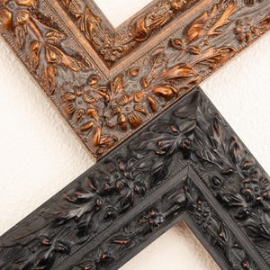 May include: Close-up of two ornate picture frame corners. One is a dark bronze color, the other is black. Both frames feature detailed floral and leaf carvings, creating a classic, vintage aesthetic. The frames are set against a white background.