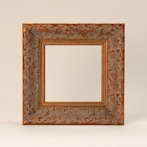 Könnte beinhalten: Quadratischer, verzierter Bilderrahmen mit Bronze-Finish. Der Rahmen weist detaillierte, erhabene Blumen- und Scrollmuster auf. Die innere Öffnung ist quadratisch und bereit für ein Foto oder Kunstwerk. Ein dekoratives Element.