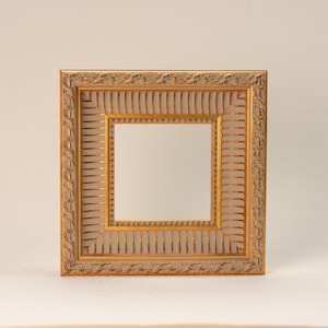 Puede incluir: Marco de fotos cuadrado y ornamentado, con acabado dorado y blanquecino. El marco presenta detallados patrones florales y líneas verticales. La abertura interior es de forma cuadrada.