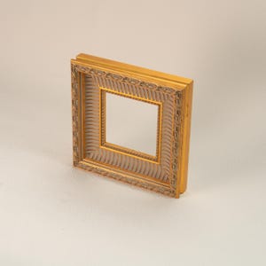 May include: A square, ornate gold picture frame with a detailed, textured design. The frame has a layered appearance with a lighter-colored inner border. The frame is empty, ready for a photo or artwork.