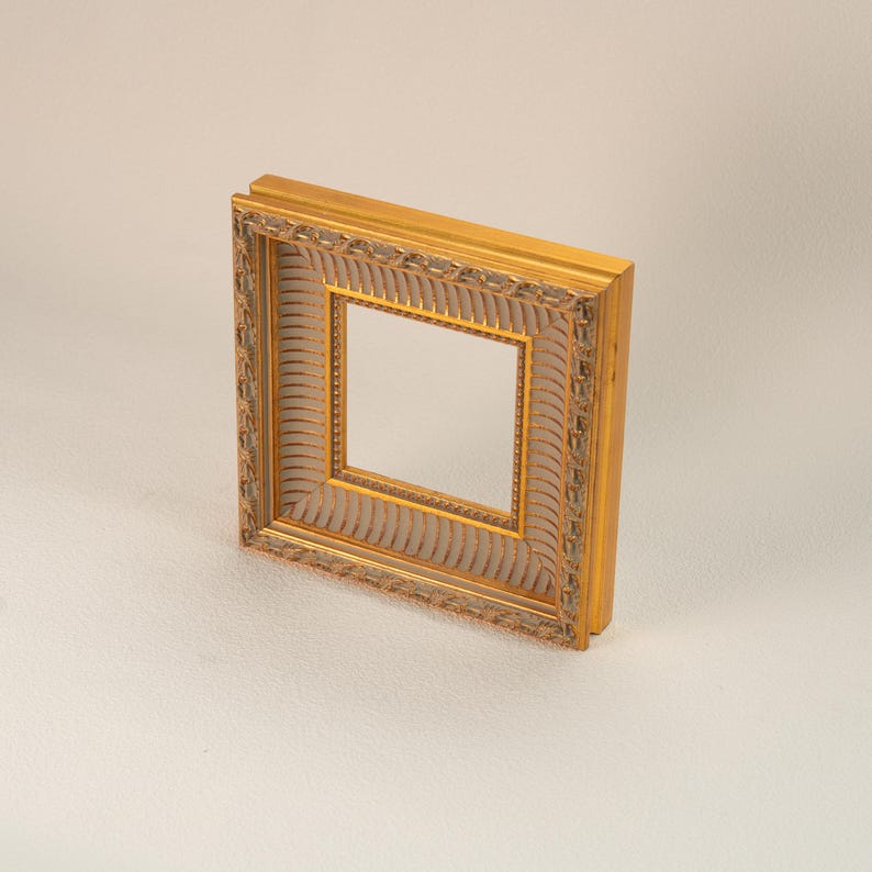 May include: A square, ornate gold picture frame with a detailed, textured design. The frame has a layered appearance with a lighter-colored inner border. The frame is empty, ready for a photo or artwork.