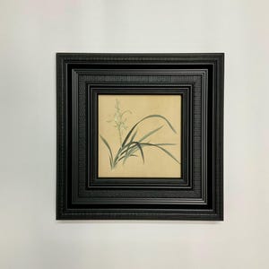 以下が含まれることがあります： ベージュの背景に、緑と白の花のイラストが描かれた、額装された植物画。 このアートワークは、黒の多層フレームに収められており、作品の美的魅力を高めています。