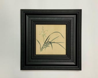 May include: A framed botanical print featuring a delicate floral illustration in shades of green and white against a beige background. The artwork is encased in a black, multi-layered frame, adding a touch of elegance to the piece.