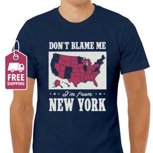 Puede incluir: Una camiseta azul marino con un gráfico blanco de un mapa de los Estados Unidos con el texto "Don't Blame Me I'm from New York".