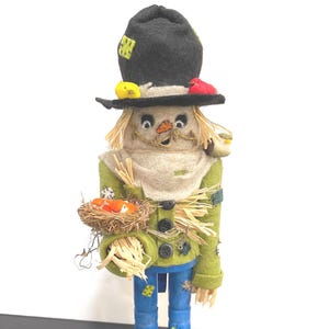 Könnte beinhalten: Eine skurrile Vogelscheuchenfigur mit einem schwarzen Hut, der mit gelben und roten Akzenten verziert ist. Die Vogelscheuche trägt eine grüne Jacke, blaue Hosen und hält ein Nest mit orangefarbenen Vögeln. Kürbisse sitzen an der Basis, perfekt für die Herbstdekoration.