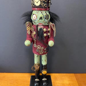 May include: A steampunk-themed nutcracker figurine. The zombie-like face is green with exposed teeth and a single eye. The uniform is burgundy with gold accents, gears, and a chain. The hat is black and burgundy, adorned with gears and a clock.