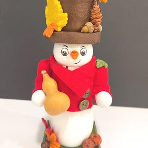 May include: A wooden snowman figurine with a white body, red jacket, and a brown top hat adorned with fall-themed decorations. The snowman holds a gourd and stands on a green base with miniature pumpkins.