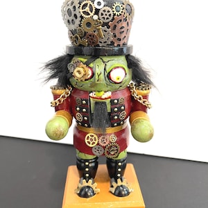 May include: A nutcracker figure with green skin, a black top hat adorned with metallic gears and keys, and a red jacket with gold chains. The figure features a monocle, black hair, a black beard, and gear embellishments on the chest and boots.
