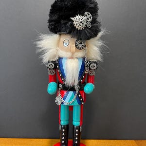 May include: A steampunk-themed nutcracker figurine. It features a black furry hat with silver gears, a monocle, and a white beard. The uniform is red, blue, and teal, with silver gear accents. The base is red and blue.