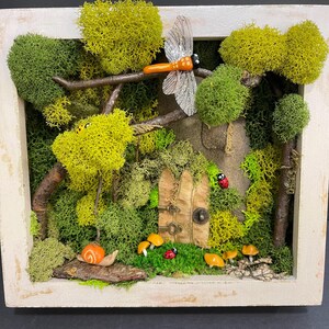 Op de afbeelding: Een grillige diorama scène in een wit omkaderde doos. De scène toont een miniatuur feeëntuin met een houten deur, mos, bomen, een libelle, paddenstoelen, een slak en lieveheersbeestjes. Het kleurenpalet omvat tinten groen, bruin, oranje en wit.