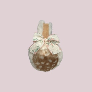 May include: A decorative ear warmer with a faux fur exterior, featuring a brown and white spotted pattern. A floral patterned bow with pink accents adorns the front. The ear warmer has a tan-colored band at the top.