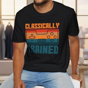 May include: Black t-shirt with the text "CLASSICALLY TRAINED" and a retro video game controller graphic in orange, red, and teal. The person is wearing blue jeans and sitting on a white surface.