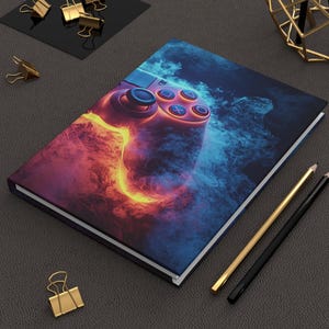 May include: A hardback journal with a striking image of a video game controller, depicted within swirling orange and blue smoke. The controller's buttons are visible. The scene is complemented by gold and black pencils and paper clips.