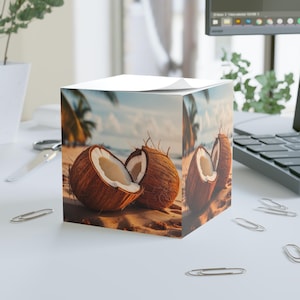 Puede incluir: Un bloc de notas cúbico con una escena de playa tropical. La imagen muestra dos cocos cortados por la mitad sobre la arena. Los cocos son marrones con interiores blancos, y el fondo muestra palmeras, el océano y un cielo azul.