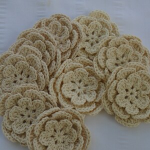 May include: A collection of cream-colored, crocheted flower appliques. Each flower has multiple layers of petals, creating a three-dimensional effect. The flowers are arranged on a white, textured surface, suggesting a craft or DIY project.