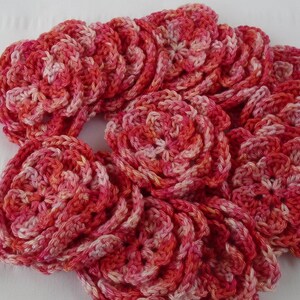 May include: A collection of crocheted flowers in shades of pink and white. The flowers are arranged in a circular pattern, creating a decorative design.