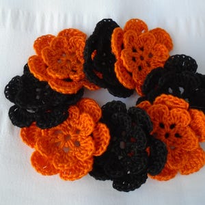 May include: A collection of handmade crochet flowers in orange and black arranged in a circular pattern. The flowers are made of yarn and have a textured appearance. The arrangement is set against a white background.