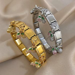 Puede incluir: Dos pulseras, una dorada y otra plateada, cada una con un diseño de eslabones rectangulares. Ambas pulseras presentan pequeños dijes con detalles de piedras preciosas rosas, verdes y transparentes, que se asemejan a cerezas. Las pulseras se exhiben sobre una superficie neutra y texturizada.