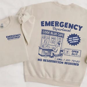 May include: Two beige sweatshirts. One has "EMERGENCY DEPARTMENT" printed on the front. The other has the same text on the front, and a graphic on the back that says "EMERGENCY Department CODE BLUE CAFE" with additional text and illustrations in blue.