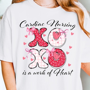 May include: White t-shirt with a graphic that reads "Cardiac Nursing XO is a work of Heart." The design features pink hearts, bows, and the letters XO in a heart shape. The shirt is a casual style.