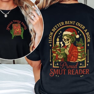 Pode incluir: T-shirt preta com um gráfico de um livro e o texto "Spread Those Pages" na frente. A parte de trás da camisa apresenta um esqueleto lendo um livro com o texto "I Look Better Bent Over a Book" e "Proud Smut Reader".