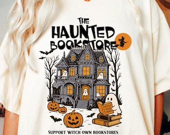 Puede incluir: Camiseta color crema con un gráfico de una casa embrujada con calabazas, murciélagos y fantasmas. El texto dice "The Haunted Bookstore" y "Support Witch Own Bookstores". Una bruja vuela en una escoba en un círculo naranja.