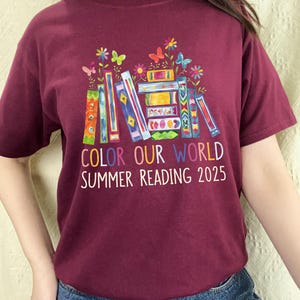 May include: Burgundy t-shirt with a colorful stack of books, butterflies, and flowers. The text reads "Color Our World Summer Reading 2025".
