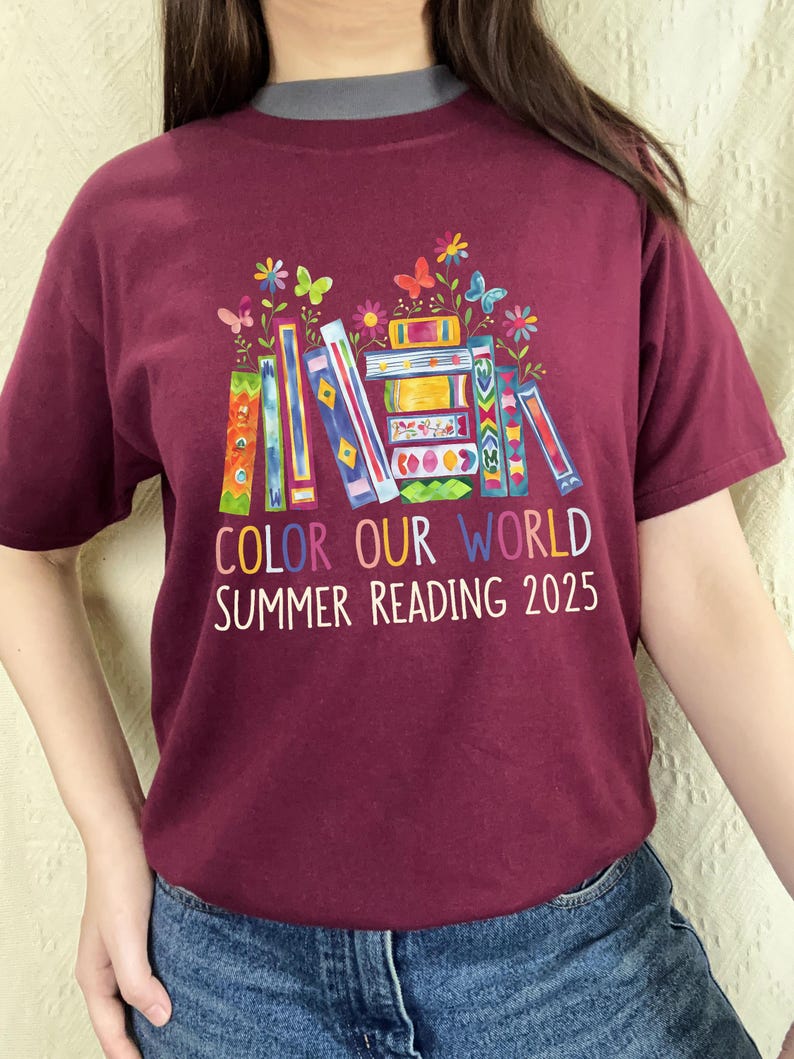 May include: Burgundy t-shirt with a colorful stack of books, butterflies, and flowers. The text reads "Color Our World Summer Reading 2025".
