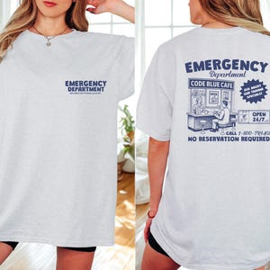 May include: A light gray t-shirt with the words "EMERGENCY DEPARTMENT" printed in navy blue on the front and back. The back of the shirt features a graphic of a cafe with the text "CODE BLUE CAFE" and "OPEN 24/7".
