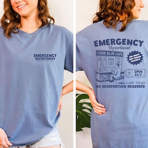 May include: A dusty blue t-shirt with the words "EMERGENCY DEPARTMENT" printed on the front. The back of the shirt features a graphic of a cafe with the text "CODE BLUE CAFE", "ASK ABOUT OUR TRAUMA SPECIALS!", and "OPEN 24/7".