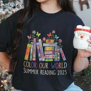 May include: Black t-shirt with a colorful illustration of stacked books and flowers. The text "Color Our World Summer Reading 2025" is printed on the shirt.