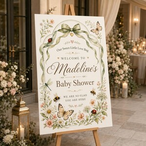 May include: A rectangular white welcome sign for a baby shower featuring a green ribbon bow at the top, floral illustrations, and insects including butterflies, ladybugs, and a dragonfly. Text reads: Our Sweet Little Love Bug Welcome To Madeline's Baby Shower We Are So Glad You Are Here.