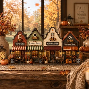 Könnte beinhalten: Ein Miniaturdorf mit vier verbundenen Gebäuden namens Maple Café, Harvest Bakery, Autumn Coffee und Cinnamon House. Jedes Gebäude verfügt über eine Ladenfront, gestreifte Markisen, eine warme Innenbeleuchtung und herbstliche Dekoration.