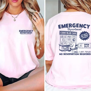 May include: A light pink t-shirt with the words "EMERGENCY DEPARTMENT" printed on the front. The back of the shirt features a graphic of a diner called "CODE BLUE CAFE" with the text "OPEN 24/7" and "NO RESERVATION REQUIRED".