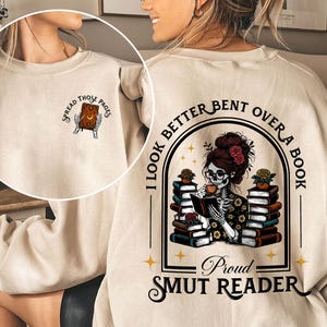 May include: A beige sweatshirt with the text "I look better bent over a book, Proud Smut Reader" and a graphic of a skeleton woman reading a book. The graphic also includes the text "Spread those pages".