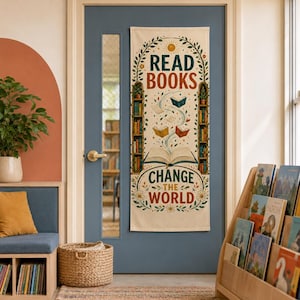 Puede incluir: Un tapiz de pared vertical de tela color crema con el texto READ BOOKS CHANGE THE WORLD. El diseño incluye ilustraciones de dos estanterías altas, un libro abierto central con páginas que se arremolinan y enredaderas florales.