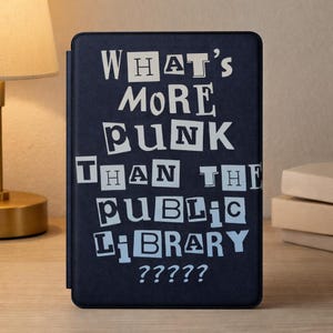 May include: A navy blue e-reader cover with the text "WHAT'S MORE PUNK THAN THE PUBLIC LIBRARY ?????" in a white, cut-out style font. The cover is rectangular and appears to be made of a textured material.