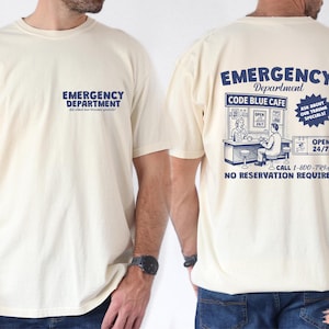 May include: Cream-colored t-shirt with the words "EMERGENCY DEPARTMENT" printed in blue on the front and back. The back of the shirt features a graphic of a cafe with the text "CODE BLUE CAFE" and "OPEN 24/7".
