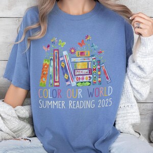 May include: A blue t-shirt with a colorful illustration of stacked books, flowers, and butterflies. The text "Color Our World Summer Reading 2025" is printed below the illustration.