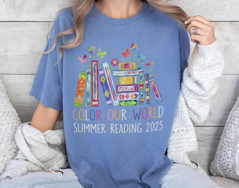 May include: A blue t-shirt with a colorful illustration of stacked books, flowers, and butterflies. The text "Color Our World Summer Reading 2025" is printed below the illustration.