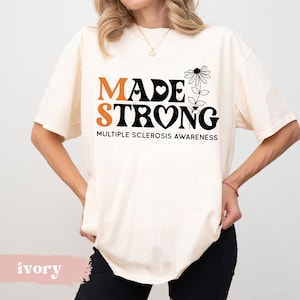May include: A white t-shirt with the text "MADE STRONG" in black and orange letters. The text "MULTIPLE SCLEROSIS AWARENESS" is printed below the main text. A black and white flower with a heart in the center is next to the text.