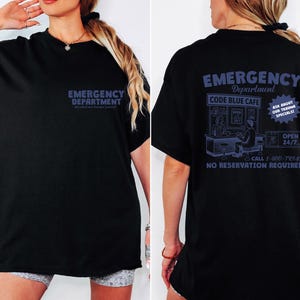 May include: Black t-shirt with blue text. The front of the shirt reads "EMERGENCY DEPARTMENT". The back of the shirt features a graphic of a cafe with the text "CODE BLUE CAFE" and "ASK ABOUT OUR TRAUMA SPECIALS!" and "OPEN 24/7".